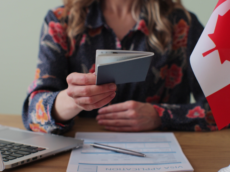 A person holding out their passport on a table beside a Canadian flag and visa application documents